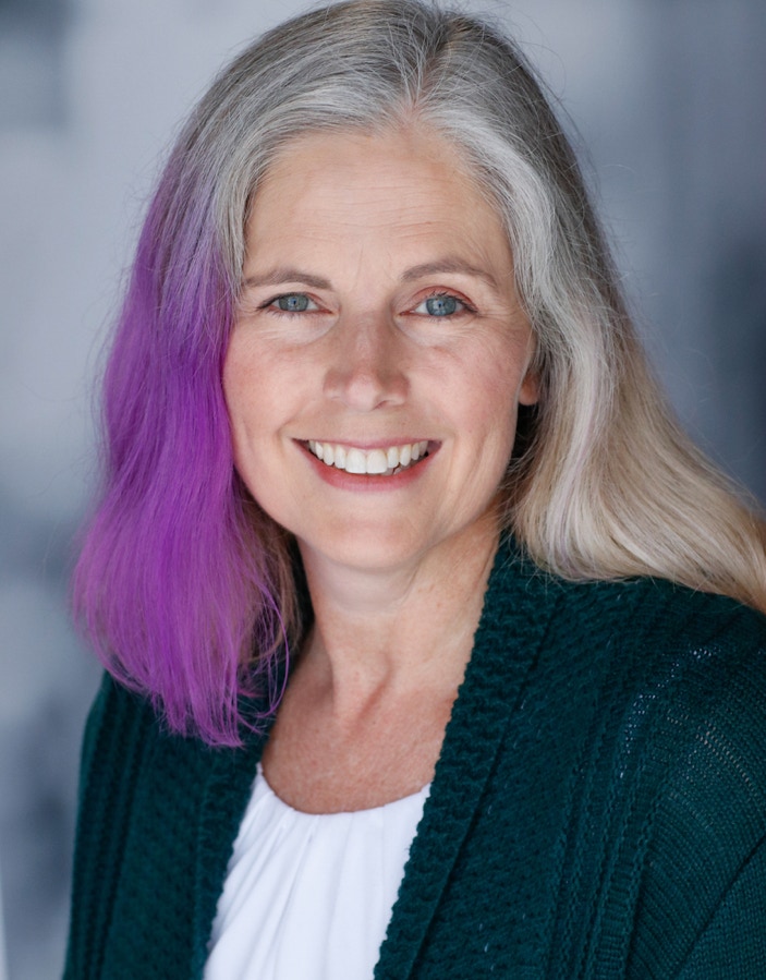 Colleen Keene vocalist is a white woman in a green sweater over a white shirt. She is smiling. Shoulder-length hair is purple on right side, silver on top, and blonde on the left.